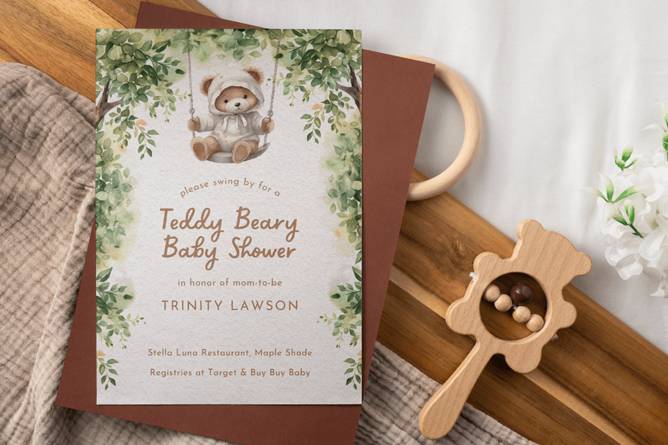 Baby shower invitation featuring a teddy bear on a swing, set on a wooden surface alongside a baby rattle.