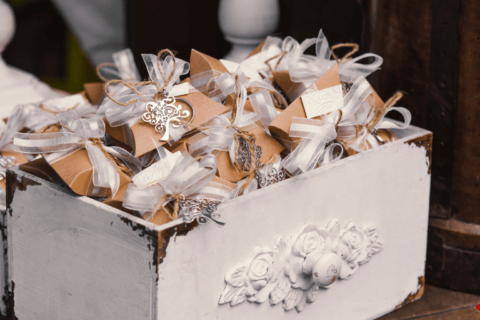 An elegantly designed ornamental white shelf, artfully utilized as a basket, brimming with an assortment of neatly wrapped party favors. Each favor is adorned with delicately tied white ribbons, adding a touch of sophistication and festive charm to the arrangement. Cover for Say thanks with 60+ Fantastic Party Favors for adults