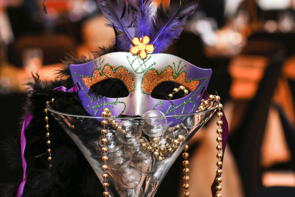 An elaborate masquerade ball mask, adorned with intricate purple and orange details, artfully placed atop a large martini glass. This creative arrangement serves as a decorative centerpiece, perfectly capturing the essence of a winter wedding with a masquerade theme