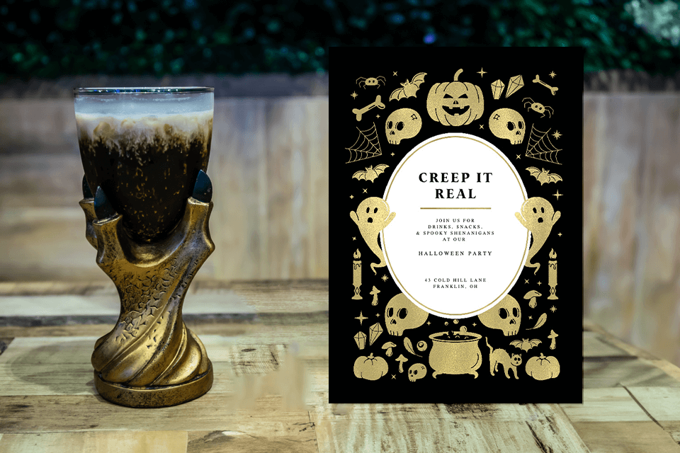 A Halloween party invitation with a black background adorned with gold illustrations of ghosts, pumpkins, skulls, bats, and spider webs. In the center, a white space contains the invitation text. The invite rests on a backdrop with a scary drinking cup as a party favor
