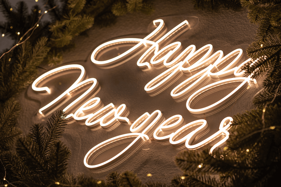 A 'Happy New Year' neon sign mounted on a wall, surrounded by pine tree branches and interwoven with string lights, creating a festive and luminous display.