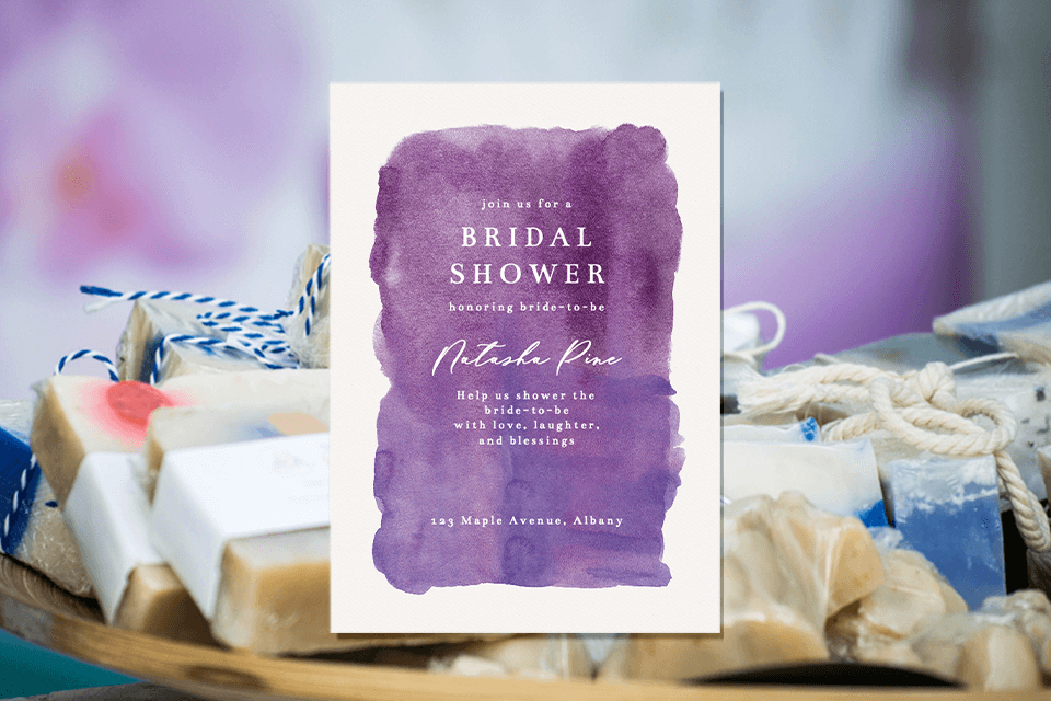 A purple watercolor bridal shower invitation, elegantly displayed on a background featuring a close-up hand made lavender soaps as party favors