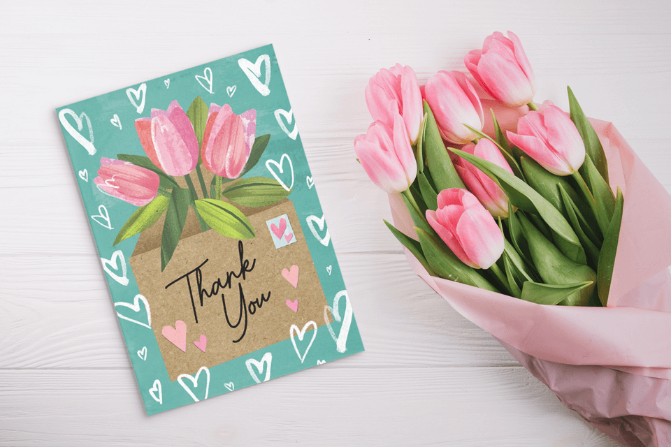 A 'Thank You' card party favor with a tulip illustration, peeking from an open envelope against a light green background adorned with white line heart drawings. The invitation rests on a wooden surface, beside a bouquet of tulips wrapped in pink paper.