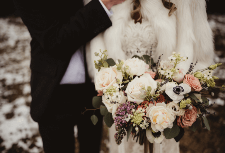 A joyful newlywed couple captured in an outdoor winter setting, epitomizing the bliss of a winter wedding. Bride and groom in the background, in the foreground the bride's elegant bouquet adds a touch of color and charm to the snowy landscape.