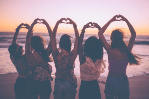 Joyful gathering of friends at the beach, backs turned to the camera, arms raised in unison to create a heart shape with their hands. Radiating unity, fun, and friendship, capturing the essence of a memorable Galentine's Day celebration.
