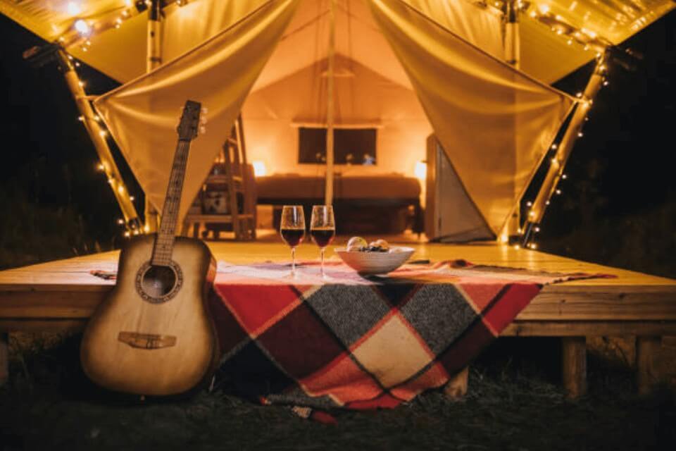 Galentine's Day Glamping Extravaganza! Our invitation showcases a luxurious glamping scene with a beautifully decorated tent adorned with twinkling string lights. Outdoors, you'll find two wine glasses, a bowl of fresh fruits, and a guitar, setting the perfect atmosphere for a memorable celebration.