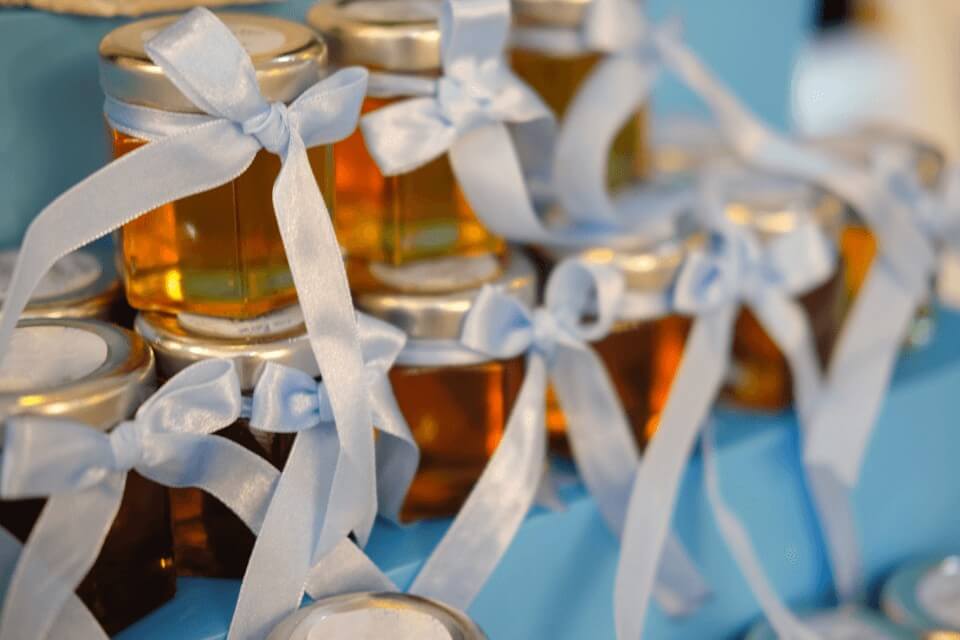 A collection of honey jars arranged neatly as party favors for an anniversary celebration. Each jar is adorned with a light blue ribbon tied in a delicate bow, adding a festive touch to the display. The jars are filled with golden honey