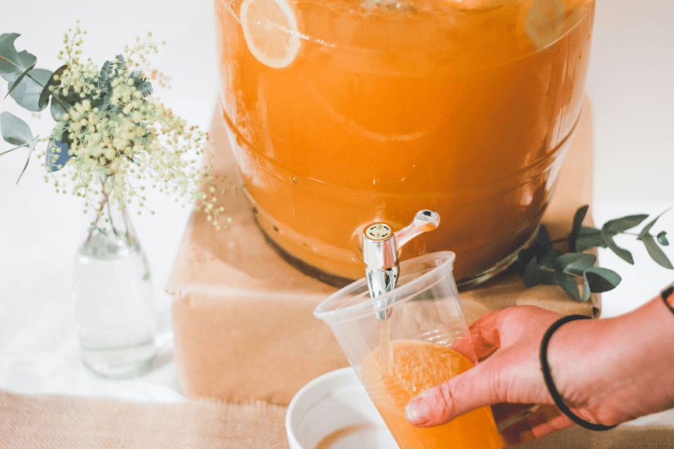 Homemade goodness on tap! A person fills a plastic cup with vibrant orange liquid from a large dispenser at a picnic birthday celebration.