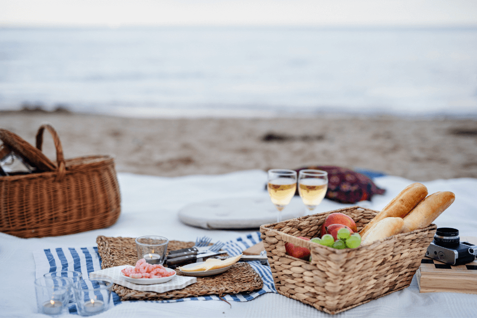  picnic blanket spread on the beach, illuminated by soft candlelight. A wicker basket brims with fresh bread and vibrant fruits, accompanied by two glasses of white wine. A camera awaits, ready to document this intimate birthday gathering.