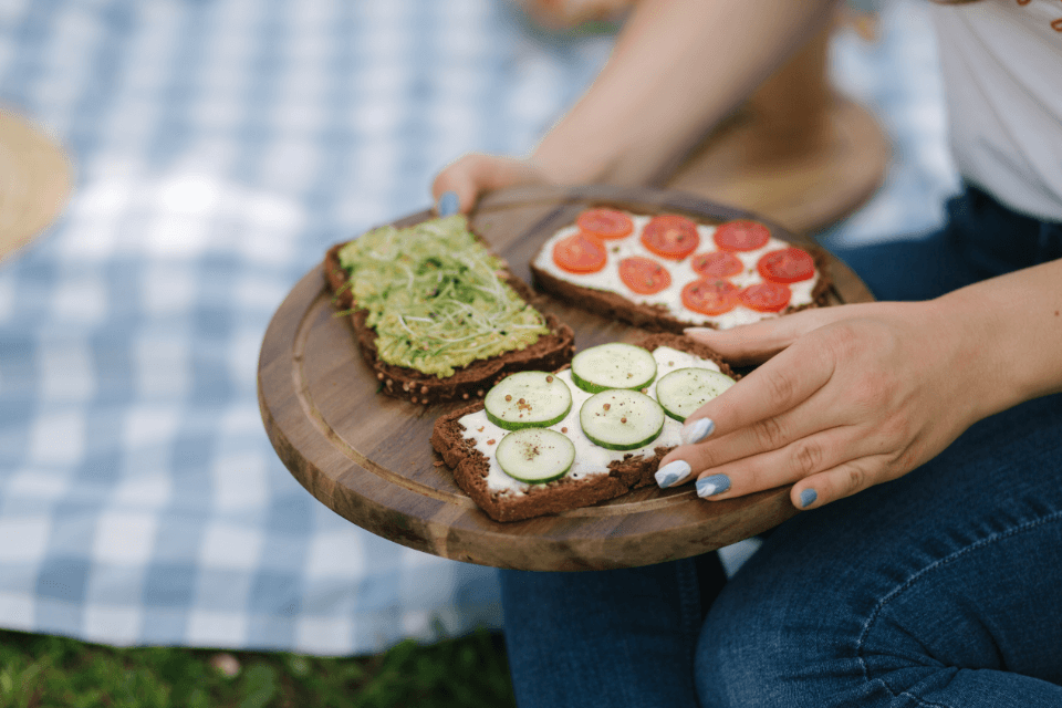 Manicured hands proudly showcase a rustic wooden tray laden with three unique sandwich creations. A vibrant picnic blanket hints at a DIY sandwich party in progress.