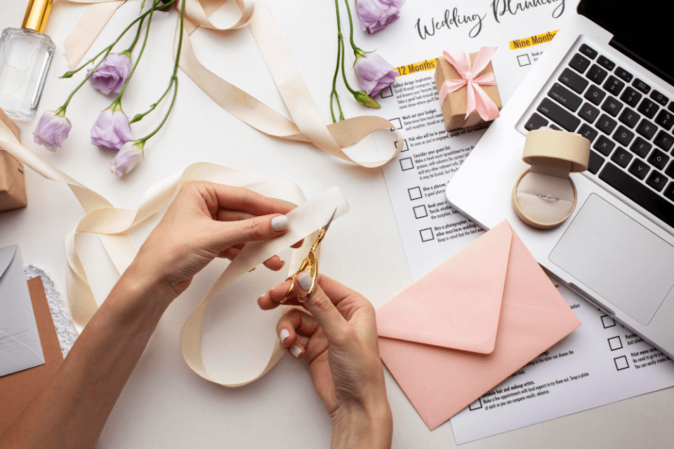 Captured in a moment of meticulous preparation, a woman delicately cuts a white ribbon with small golden scissors, crafting invitations for the bridal party. Beside her, a laptop stands ready for organization, while a ring box proudly displays its contents alongside a scattering of flowers
