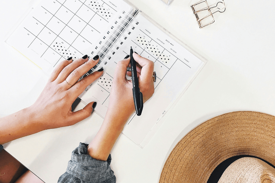 A woman's hand, grasping a black pen, meticulously plans a bridal shower on a calendar notebook. Adjacent, a straw hat hints at sunny, relaxed vibes