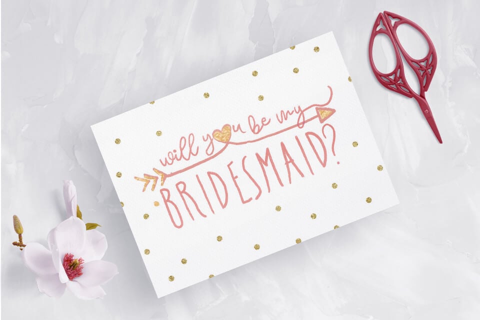 Will you be my bridesmaid?" card exuding glamour with golden dots accentuating vibrant pink text. Alongside rests a delicate flower and a pair of scissors