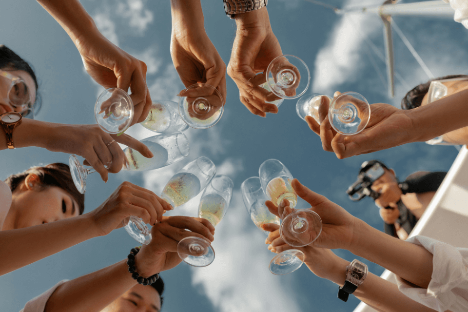 Bridal party shower: joyful women cheer with champagne glasses, captured from a dynamic low-angle perspective against a backdrop of radiant blue skies, infusing the moment with celebratory energy and sunny optimism