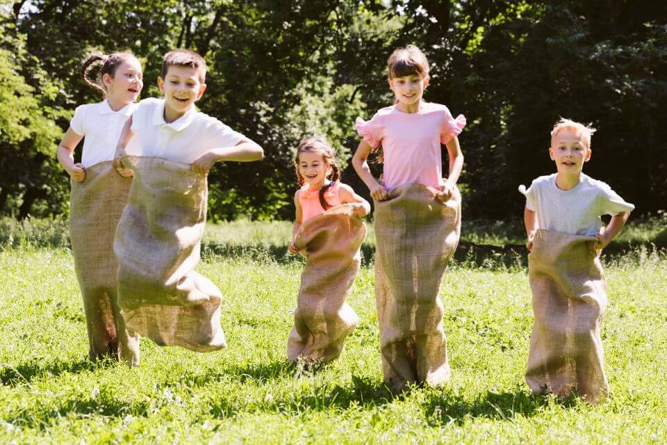 Young children engaged in a lively preschool graduation party activity, participating in a burlap sack race contest outdoors. Their joyous expressions and energetic movements capture the spirited fun of this classic game