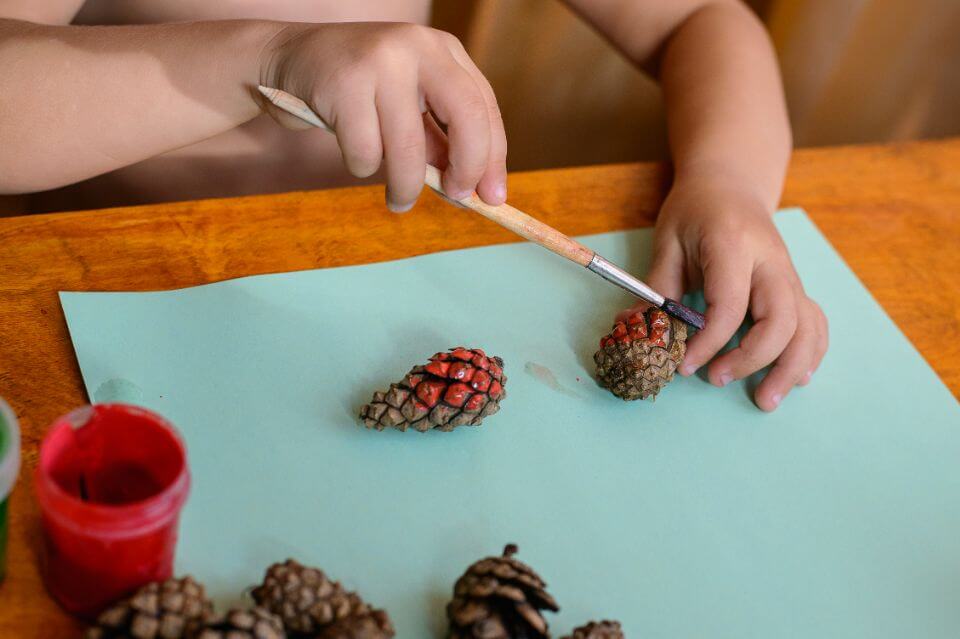 Young child's hands delicately painting small pinecones with bright red color, engaging in a creative activity as part of a preschool graduation celebration.