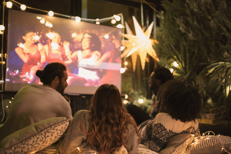 movie night outdoor friends party backyard Friends enjoying a cozy backyard movie night with pillows, blankets, string lights, and a big outdoor screen showing a film.