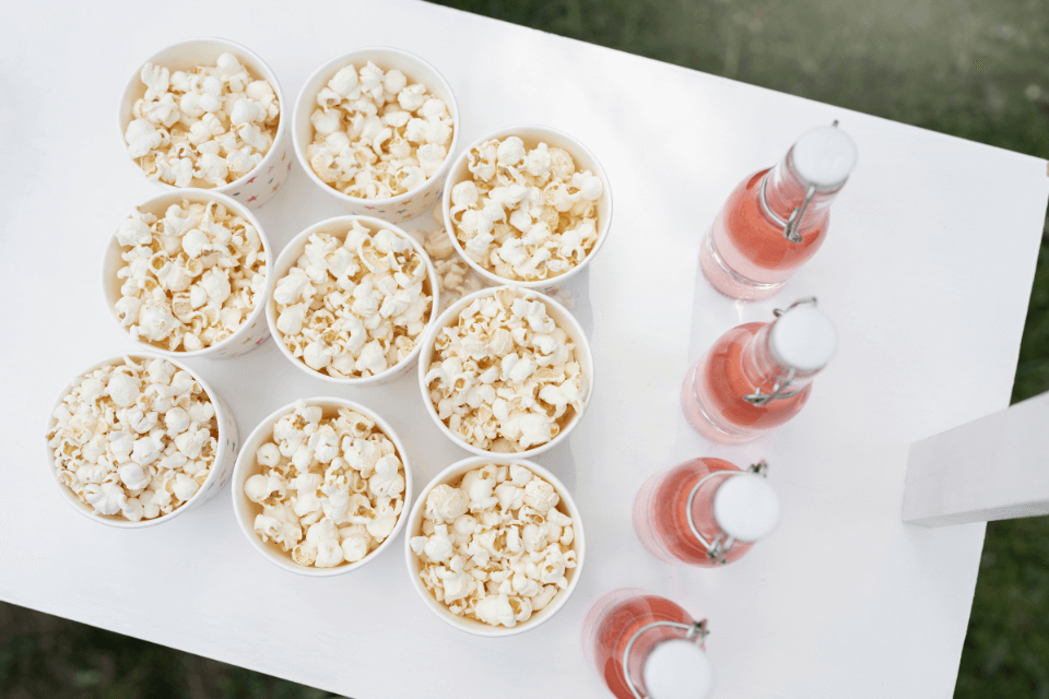 movie night party popcorn snack bar Top view of a movie night snack setup with paper cups filled with popcorn and glass bottles of pink soda neatly arranged on a white table outdoors.