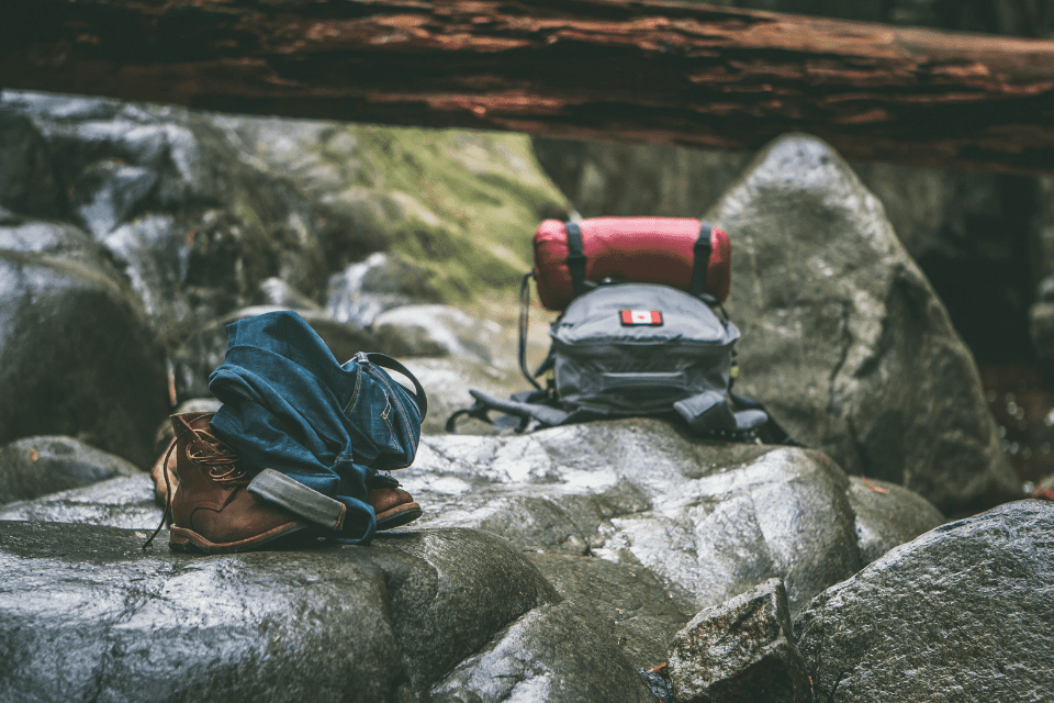 birthday hiking 40th adventure ideas Outdoor scene with hiking gear placed on wet rocks, including brown boots, rolled-up jeans, and a gray backpack with a red sleeping bag attached. The setting is a forested rocky area, suggesting a hiking or adventure theme.