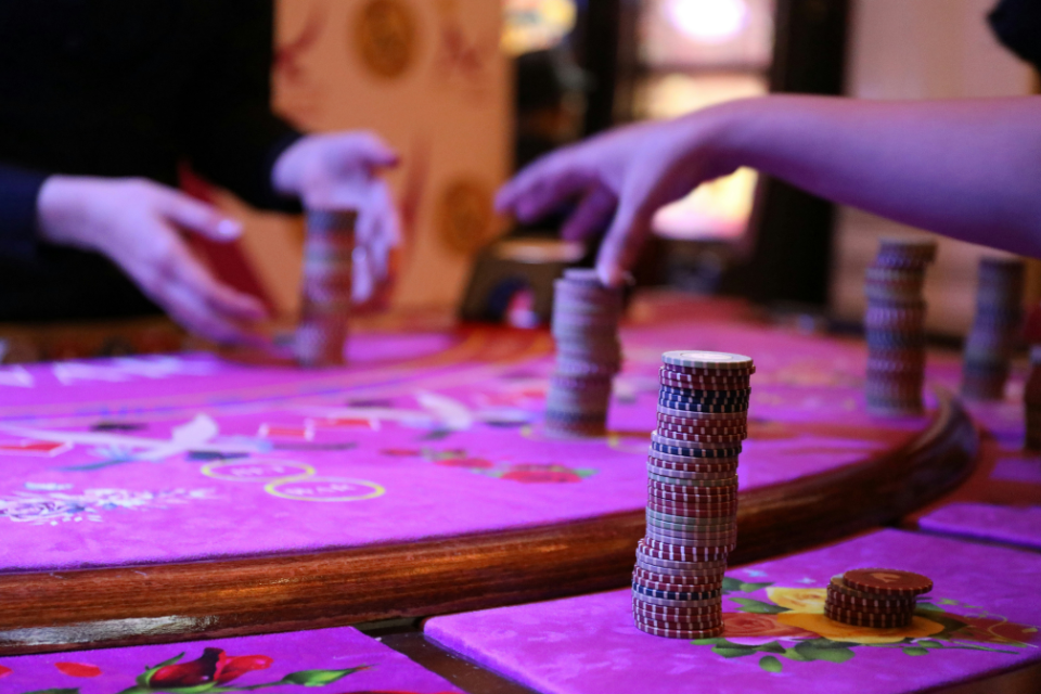 casino games 40th birthday themes Casino table scene with stacks of poker chips on a bright purple felt surface, while two people reach in to place or move chips under warm, colorful lighting.