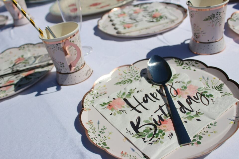 happy birthday outdoor set up Outdoor birthday table setup with floral paper plates, matching cups, striped straws, and a napkin that reads ‘Happy Birthday,’ with a spoon resting on top.