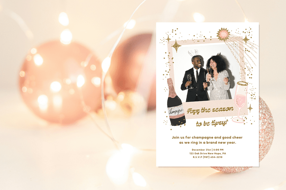 new-TIS-THE-SEASON-NEW-YEAR-DECOR-INVITE Festive New Year’s Eve invitation with a playful design featuring a framed photo of a smiling couple dressed in formal attire holding champagne glasses. Surrounding the frame are illustrated gold stars, sparkling accents, and pink champagne-themed graphics, including a bottle and flute. The text reads ‘Fizz the season to be tipsy!’ against a soft, glowing background with blurred holiday ornaments.