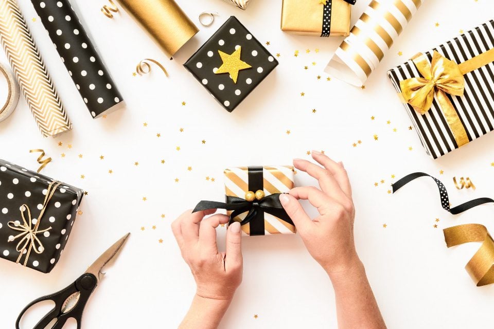 party favors new year gift wrapping Hands wrapping a small gift box with black and gold ribbon, surrounded by various wrapped presents, scissors, ribbon rolls, and gold star confetti on a white tabletop. The scene features a black, gold, and white color palette with polka-dot and striped wrapping paper.