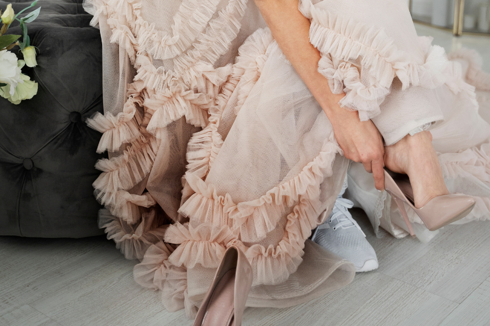 1 quinceañera changing shoes girl 15th birthday (1) Close-up of a quinceañera wearing a blush ruffled tulle gown while changing from high heels into white sneakers, highlighting the contrast between formal dress and comfortable shoes.