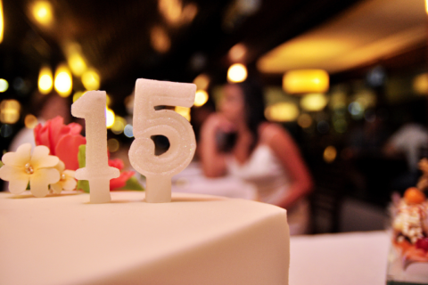 Close-up of a quinceañera cake topped with glittery number 15 candles and delicate flowers, set against a softly blurred indoor celebration with warm golden lights.