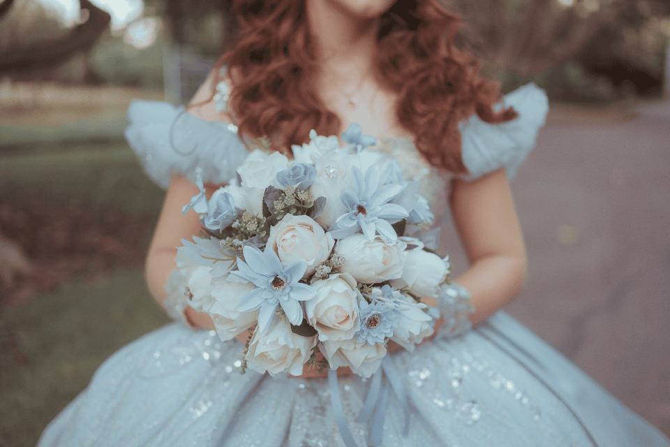 quinceañera-girl dress gown birthday festive traditional (1) Close-up of a girl in a light blue quinceañera gown holding a bouquet of pale blue and white flowers, with soft sparkles on the dress and a blurred outdoor background.