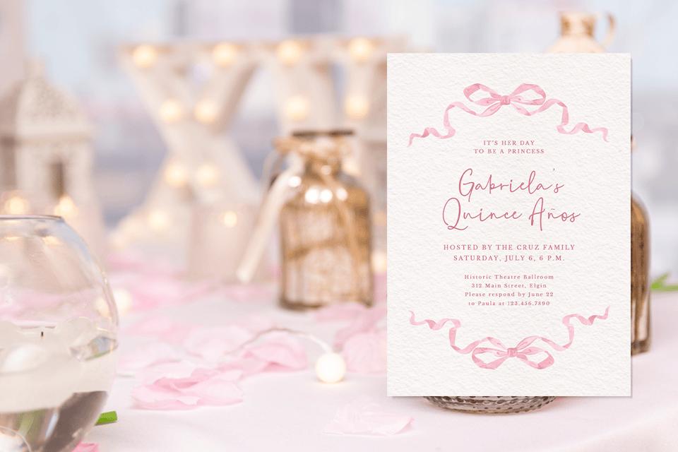 quinceañera-pink-bow-invite (1) Pink quinceañera invitation with hand-drawn ribbon bows and elegant script, displayed on a softly styled table with glass jars, fairy lights, and blush rose petals.