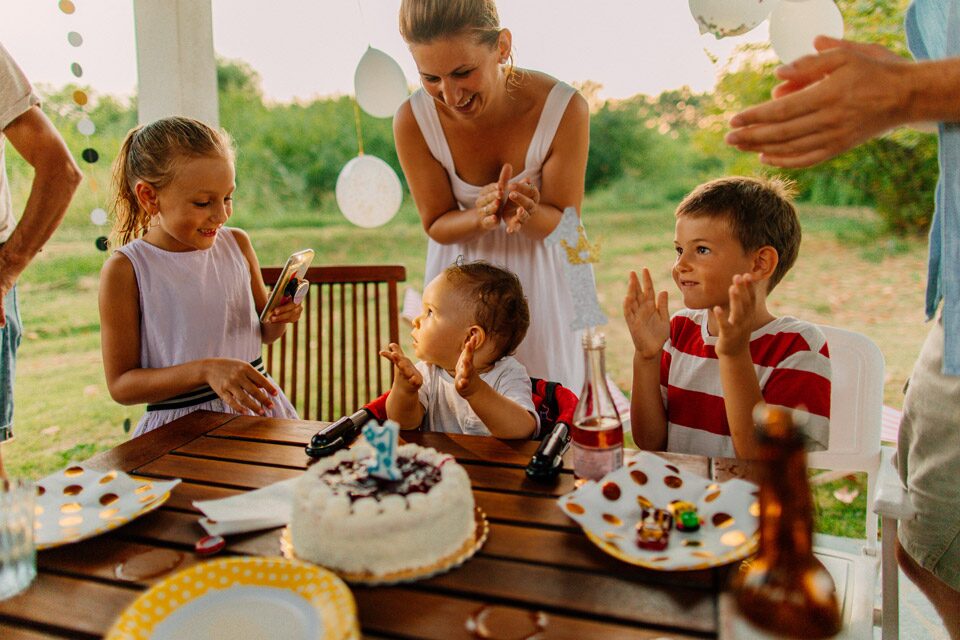 1 birthday-boy-1st-party-cake (1) Outdoor first birthday celebration with a baby sitting at a wooden table in front of a small cake with a number one candle, surrounded by smiling family members clapping and enjoying the party.