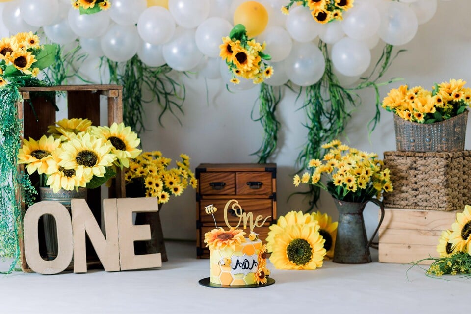 1 birthday-boy-one-sunflowers-sunny-birthday-decor Sunflower-themed first birthday party setup featuring a small honeycomb cake with “one” topper, large wooden “ONE” letters, white and yellow balloon garland, and bright sunflower arrangements in rustic crates and baskets.