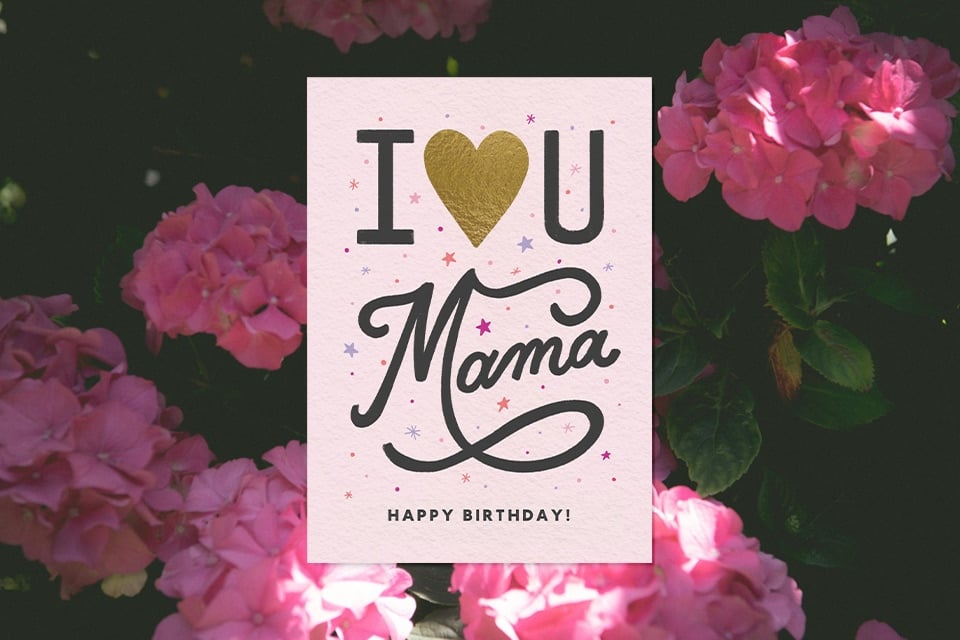 I-LOVE-YOU-MAMA-birthday-card (1) “I love you Mama” birthday card with hand-lettered text and a gold heart on a soft pink background, surrounded by blooming pink flowers in natural light.