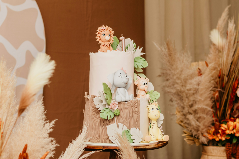Two-tier jungle-themed first birthday cake decorated with fondant animals including a lion, elephant, monkey, and giraffe, surrounded by soft neutral pampas grass décor in a warm-toned setting.