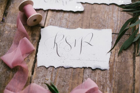 Handwritten “RSVP” calligraphy on textured paper placed on a rustic wooden table, styled with pink ribbon and greenery for an elegant invitation or event planning concept.