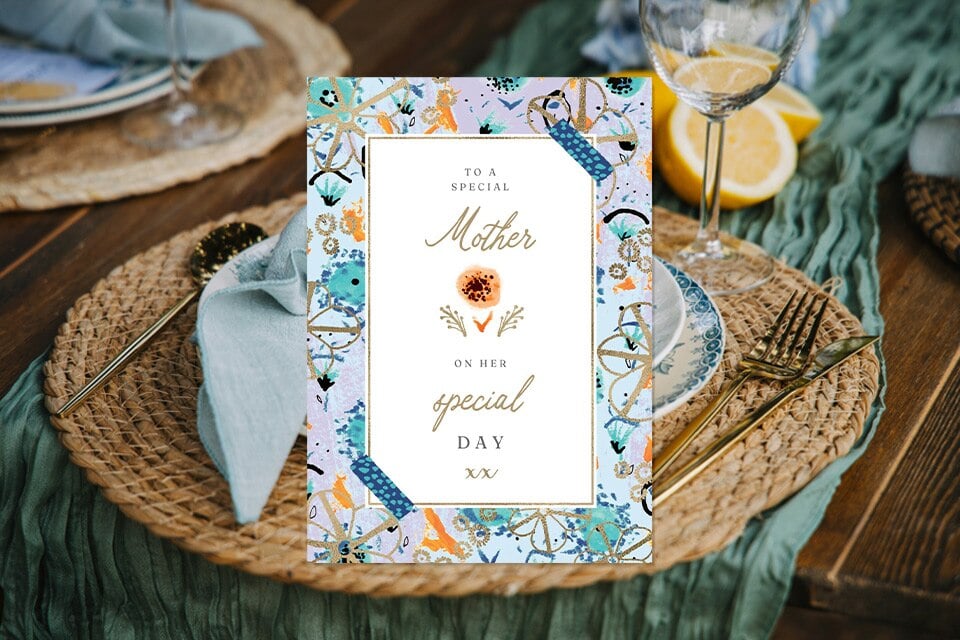 to-a-special-mother-birthday-card (1) Elegant birthday card reading “To a Special Mother on her special day,” featuring a floral and botanical border in soft blue and gold tones, placed on a rustic table setting with woven placemats, gold cutlery, linen napkins, and a wine glass with lemon slices nearby.
