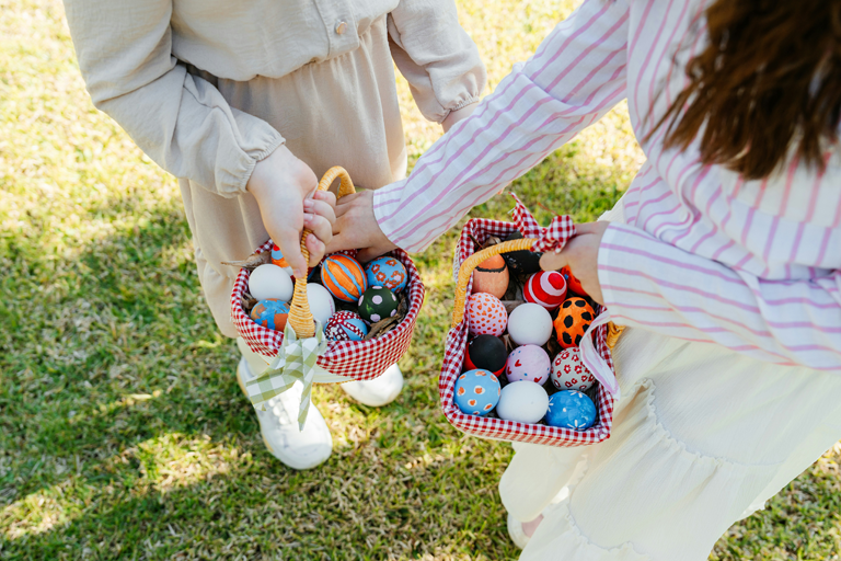 Two children holding baskets filled with colorful decorated Easter eggs while standing on grass, reaching toward each other during an outdoor egg hunt.