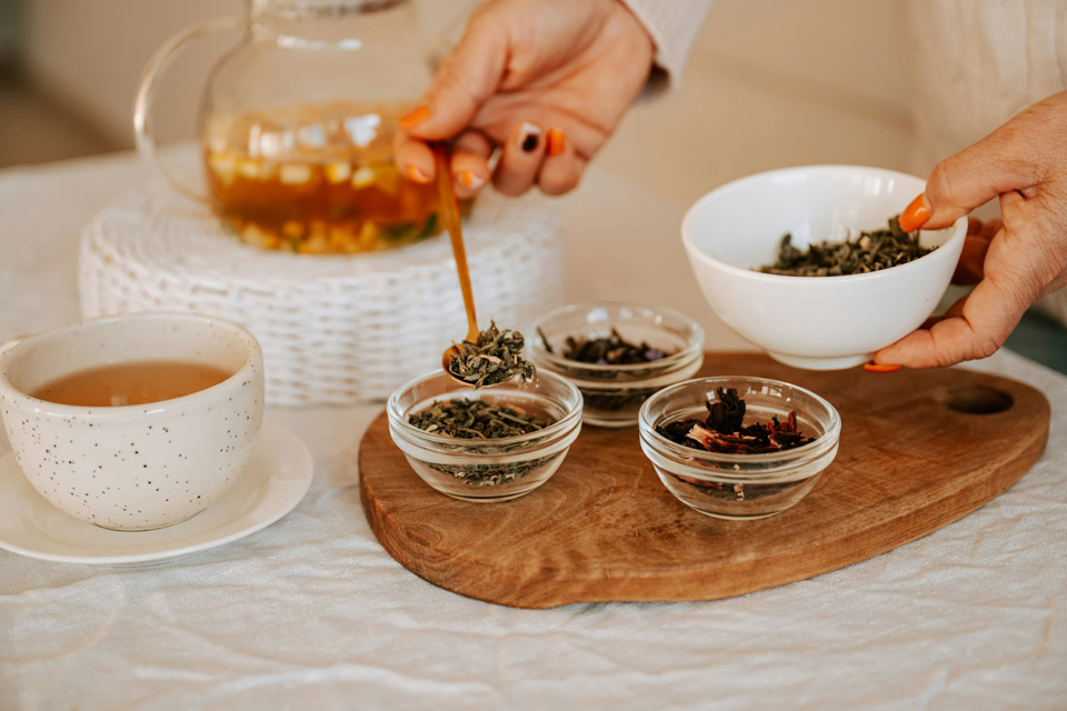 tea-party-tea-variety Hands selecting loose-leaf tea from small glass bowls on a wooden tray beside a teacup and glass teapot, showcasing a variety of teas for a tea party or tasting.