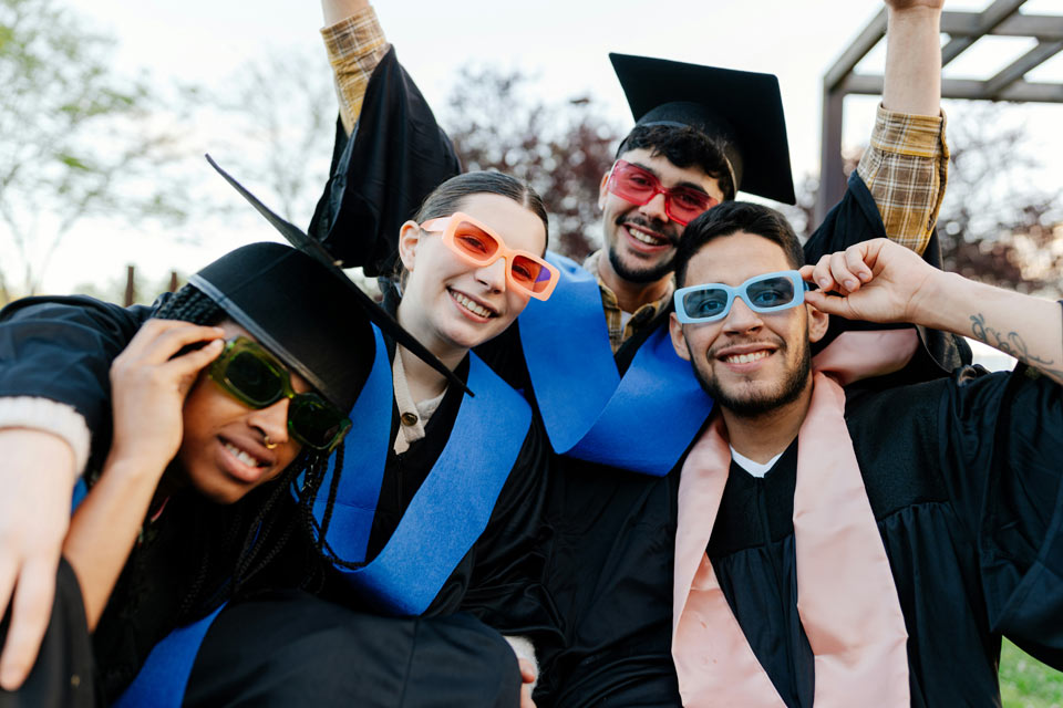 students-graduates-high-school-propr Group of graduates in caps and gowns posing together outdoors, smiling and wearing colorful sunglasses.