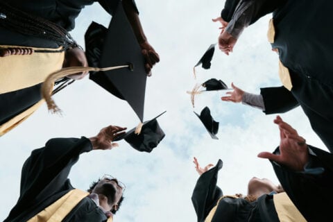 Graduates in caps and gowns tossing their mortarboards into the air, viewed from below against a bright sky.